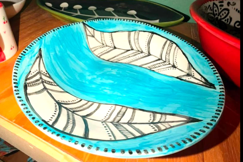 A fired plate painted turquoise with two black and white patterned feathers and a dotted rim