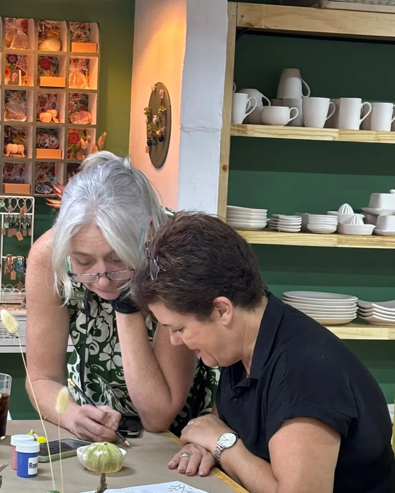 Katy leaning over the table to help a customer with a design, shelves of white bisque mugs and plates behind them