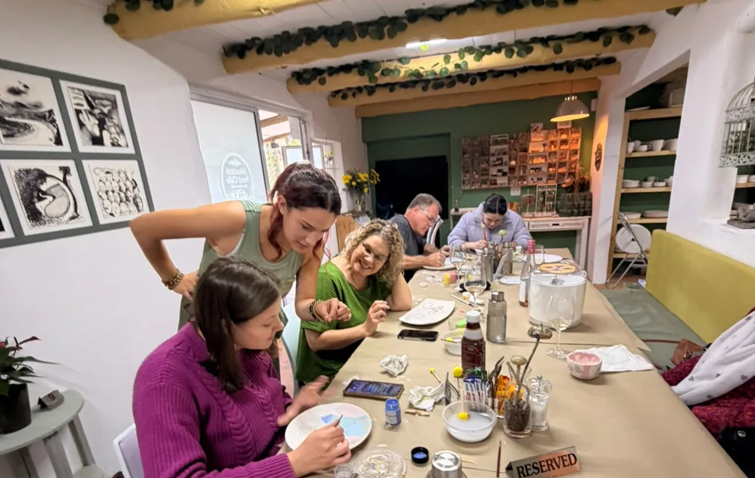 A group painting pottery along the long table under the leaf-wrapped beams, Angie helping one of them